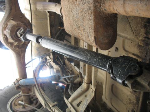 1987 Ford Bronco - Underside view of a vehicle's drivetrain, showing a driveshaft connecting to the rear differential. Leaf springs and a shock absorber are a…