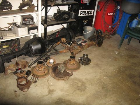 1987 Ford Bronco - A collection of automotive axle and brake parts are laid out on a concrete floor in a garage setting. Visible parts include a large rear ax…