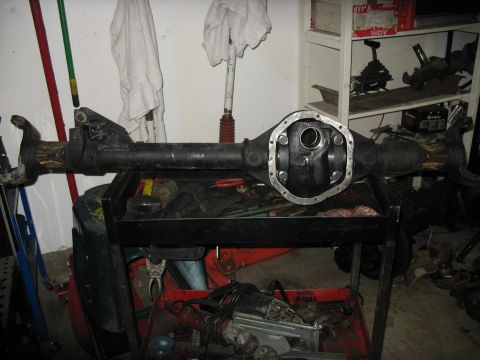 1987 Ford Bronco - A bare front axle housing rests across a rolling tool cart in a garage, with the differential cover removed to expose the empty pumpkin and…
