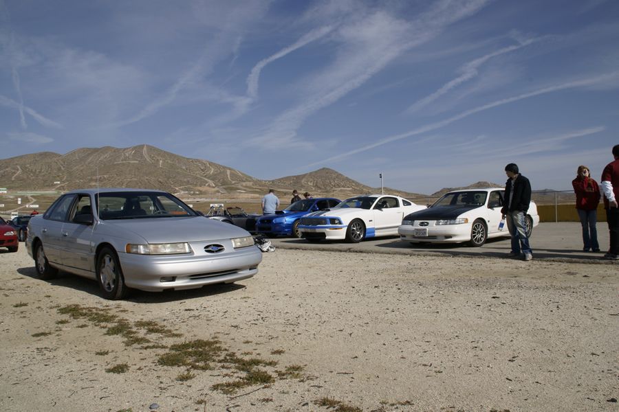 Silver 1993 Ford Taurus sedan parked on dirt lot with other cars and people.