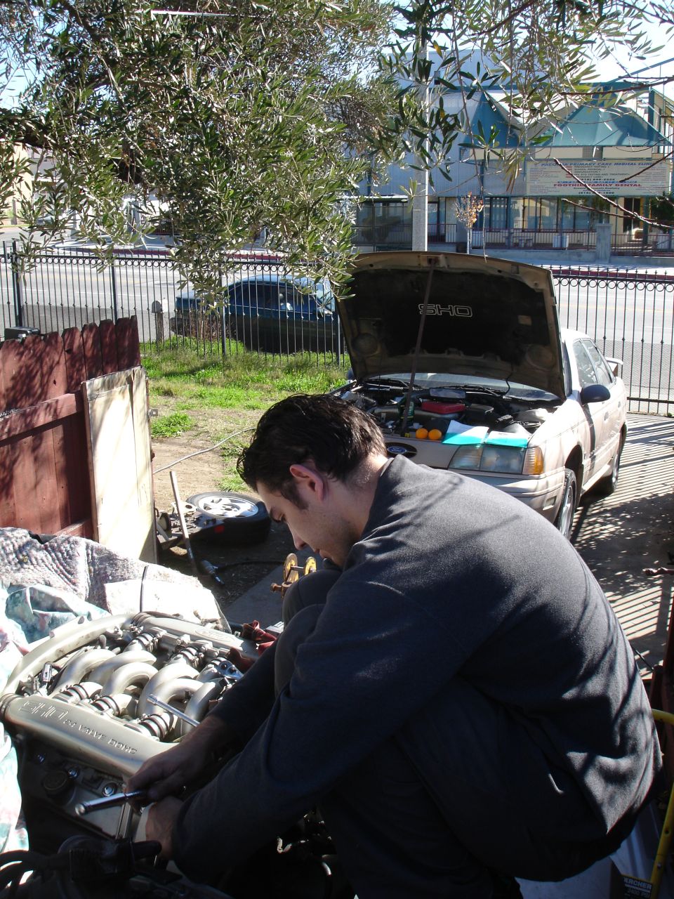 Man wrenching on a pulled 24-valve V6 engine with a tan SHO sedan hood-up behind him