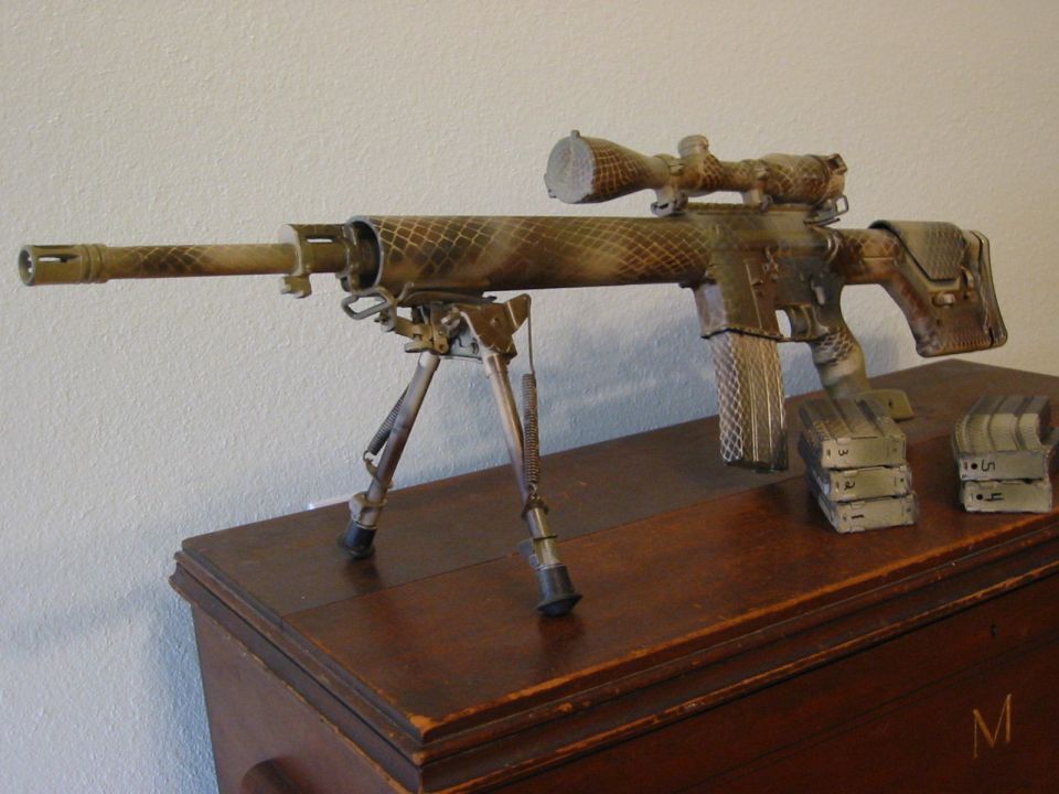 Camouflage sniper rifle with scope, bipod, and magazines on a wooden table.