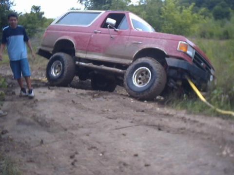 1989 Ford Bronco - A red SUV with a white roof is stuck in the mud, with a yellow tow strap attached to its front bumper.