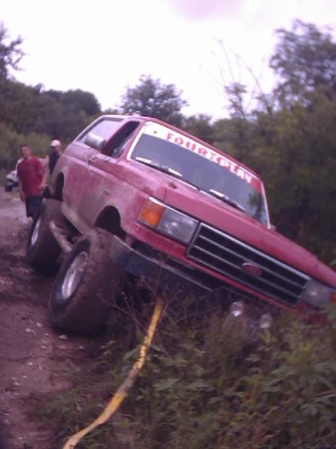 1989 Ford Bronco - A red 1989 Ford Bronco is stuck in the mud, with a yellow strap attached and a 'FOURPLAY' sticker on the windshield.