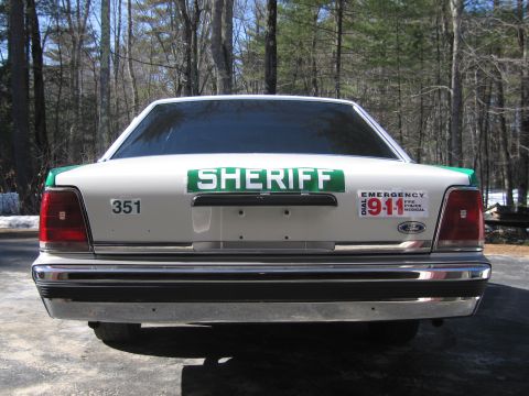 1991 Ford LTD Crown Victoria - Rear view of a white-and-green sedan marked SHERIFF with a 351 decal and DIAL 911 EMERGENCY sticker, parked on pavement by woods.