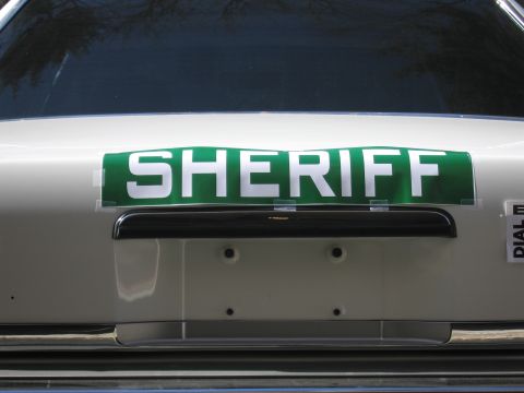 1991 Ford LTD Crown Victoria - Close-up of a green SHERIFF placard mounted on the trunk lid of a light-colored sedan above the rear license plate area.
