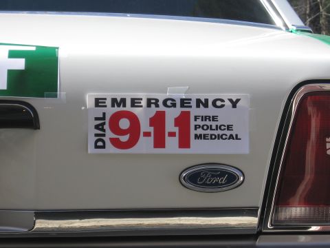 1991 Ford LTD Crown Victoria - Close-up of a DIAL 911 EMERGENCY FIRE POLICE MEDICAL decal on a white trunk beside a Ford oval emblem and the taillight.