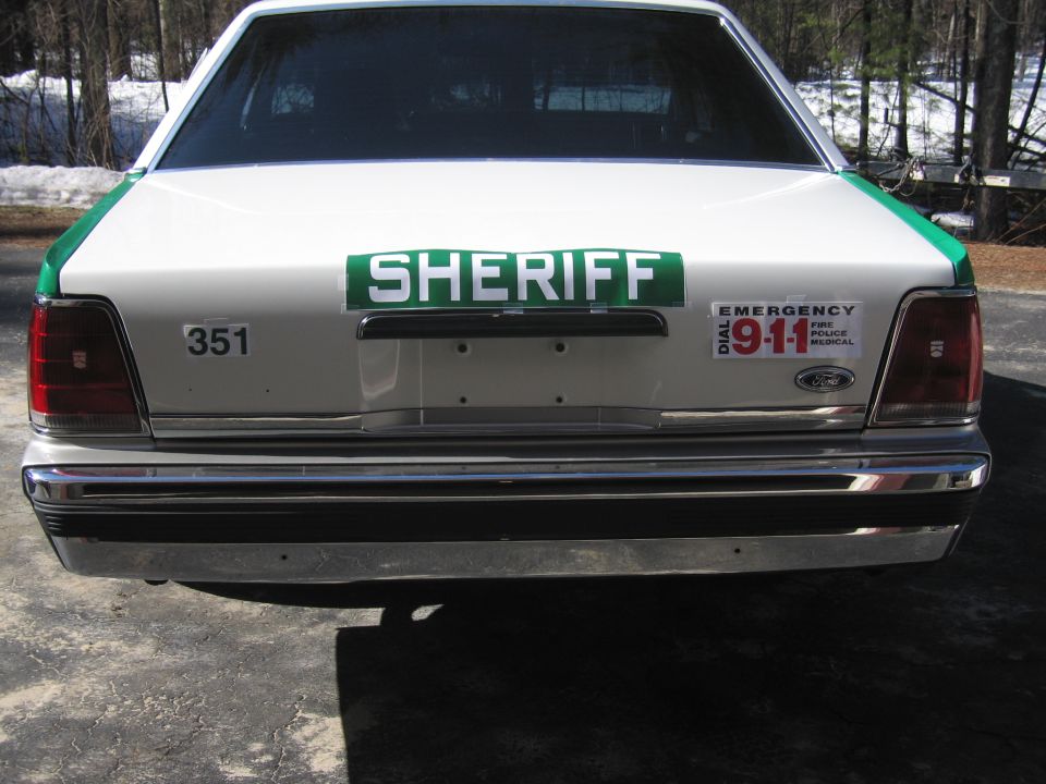 Rear of white sedan with green SHERIFF placard and 911 decal, snow in background