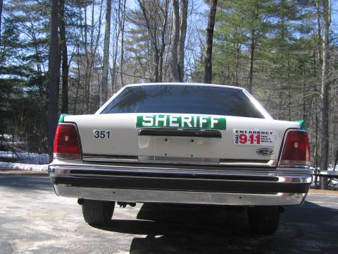 1991 Ford LTD Crown Victoria - Rear three-quarter rear view of a white-and-green SHERIFF sedan with 351 and DIAL 911 decals parked on a paved driveway by woods.