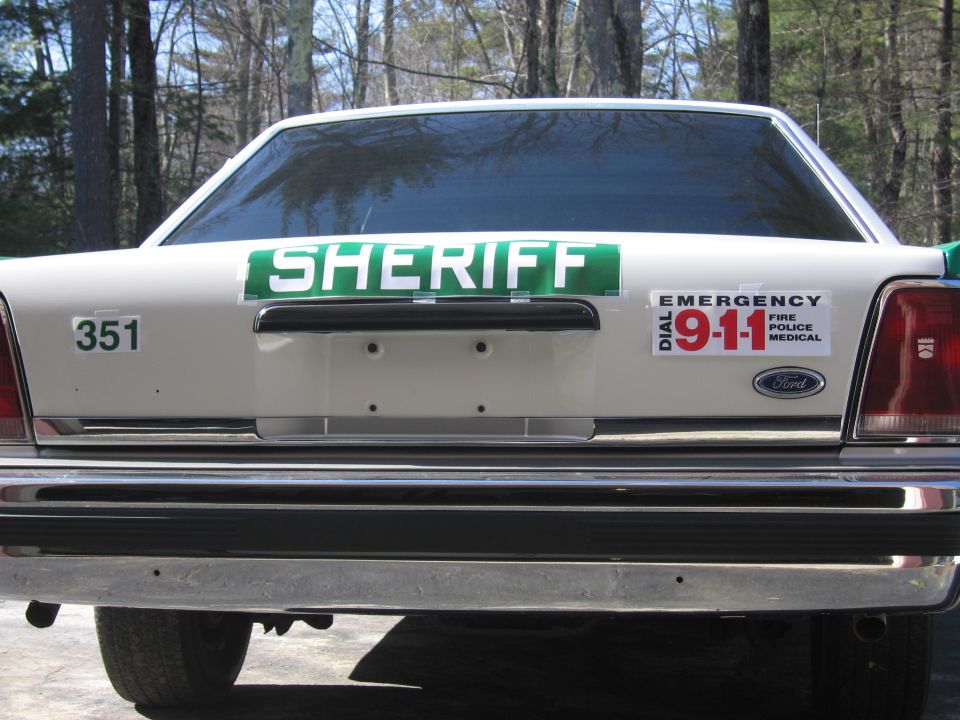 Rear of white SHERIFF sedan with green accents and DIAL 911 decal
