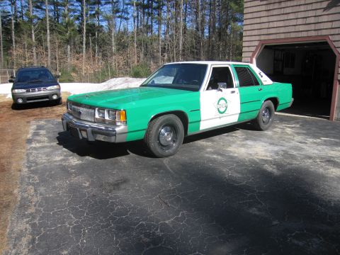 1991 Ford LTD Crown Victoria - Front three-quarter view of a green-and-white Ford SHERIFF sedan parked on a cracked driveway beside an open garage bay and a dark wagon.