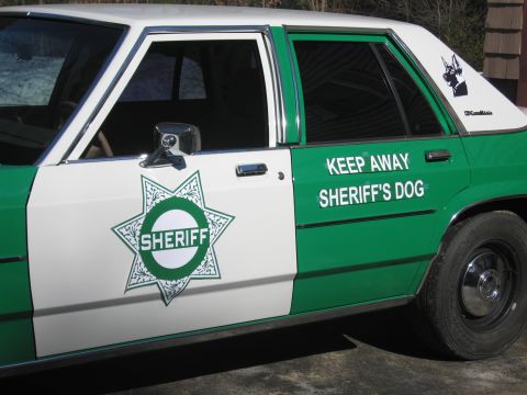 1991 Ford LTD Crown Victoria - Driver-side view of a green-and-white SHERIFF sedan with a star badge, spotlight, and KEEP AWAY SHERIFF'S DOG decal on the rear door.