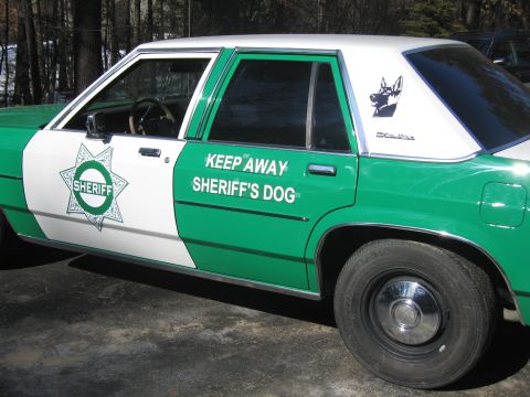 1991 Ford LTD Crown Victoria - Driver-side view of a green-and-white SHERIFF sedan showing the star badge, KEEP AWAY SHERIFF'S DOG decal and a dog silhouette graphic on t…