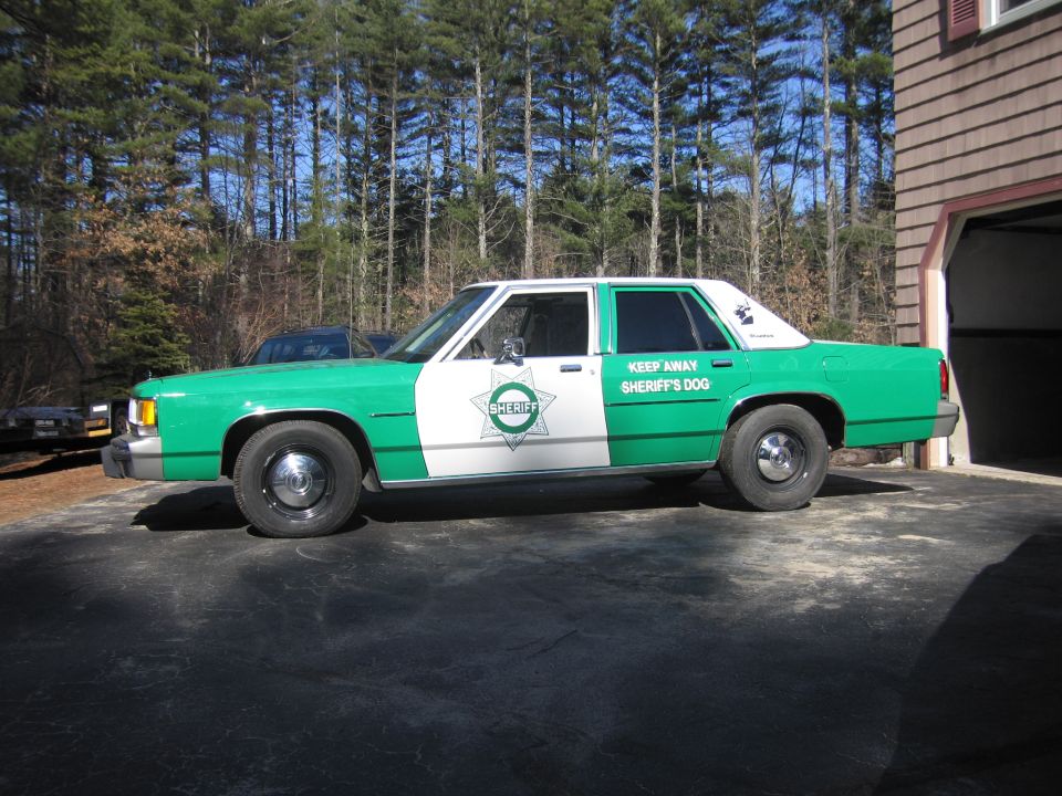 Front driver-side view of green-and-white SHERIFF sedan parked by a garage