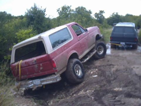 1989 Ford Bronco - A red SUV is stuck in the mud, with another vehicle behind it on a dirt road.