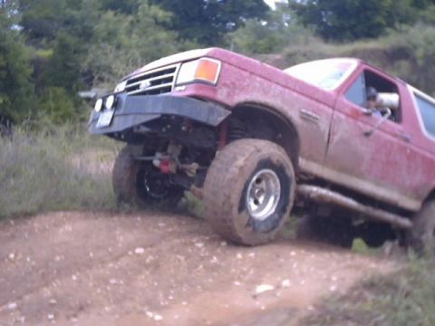 1989 Ford Bronco - A red Ford Bronco navigates a muddy off-road course, showing its lifted suspension and aggressive tires.