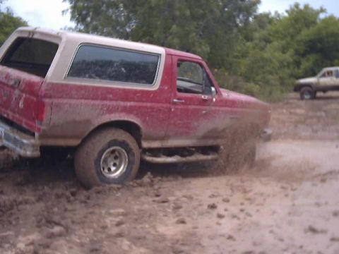 1989 Ford Bronco - A red 1989 Ford Bronco navigates a muddy off-road trail, kicking up spray as it goes.