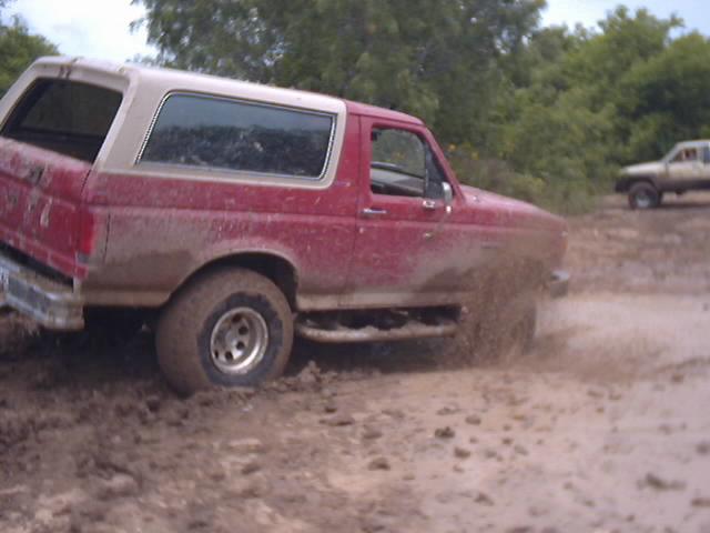 Red 1989 Ford Bronco driving through mud, splashing water. Another truck is visible in the background