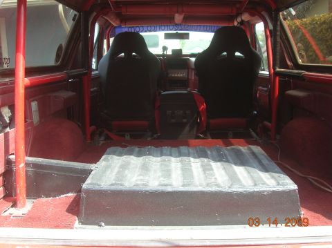 1985 Ford Bronco - The interior of a red 1985 Ford Bronco shows two black racing seats and a large black storage box with a ribbed top in the rear cargo area.