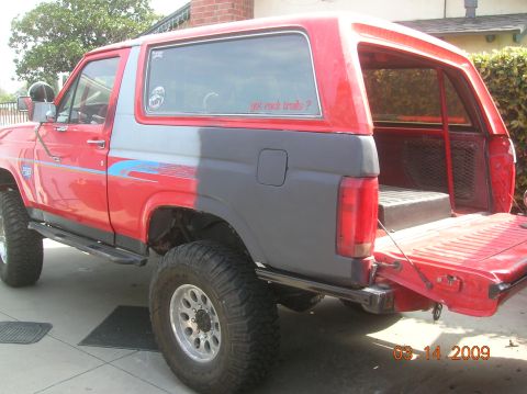 1985 Ford Bronco - The rear passenger side of a red and gray Ford Bronco is shown with the tailgate down. The vehicle has large off-road tires and a custom si…