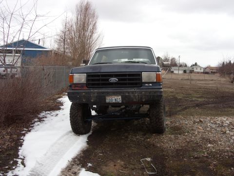 1988 Ford Bronco - Head-on front view of a lifted dark blue Ford Bronco parked on a muddy lot with snow still on the ground, showing the blue Ford oval grille…