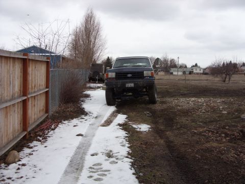 1988 Ford Bronco - A dark blue Ford Bronco is parked on a dirt and snow-covered driveway next to a wooden fence. The vehicle is facing forward, with its front…