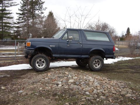 1988 Ford Bronco - A dark blue, lifted Ford Bronco is parked on a rocky incline with patches of snow on the ground. The side profile of the SUV shows its off-…