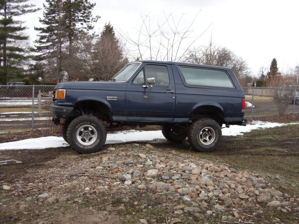 Dark blue lifted 1988 Ford Bronco, side view, parked on rocks with snow.