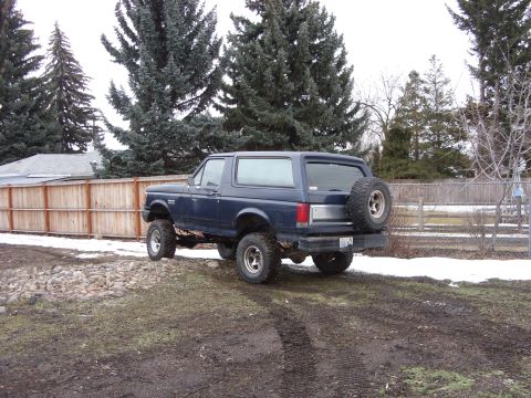 1988 Ford Bronco - Rear three-quarter view of a lifted dark blue full-size Ford Bronco with hardtop, sitting on muddy grass beside patches of snow and a wood…