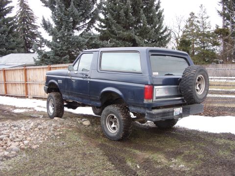 1988 Ford Bronco - A dark blue Ford Bronco is parked on a gravel and dirt lot with snow patches visible. The SUV is shown from a rear three-quarter angle, hig…