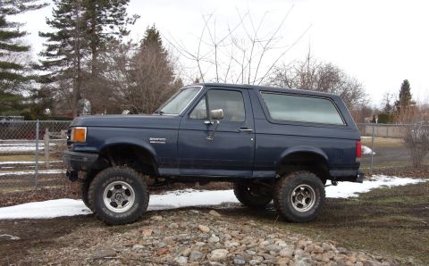 1988 Ford Bronco - A dark blue 1988 Ford Bronco is parked on a gravelly area with patches of snow and mud. The SUV is shown from the side with its front and r…