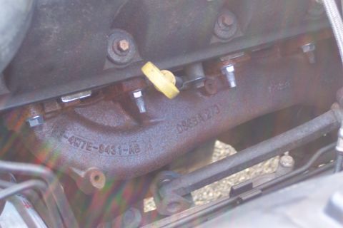 2001 Ford Crown Victoria - A close-up view of a rusty exhaust manifold with visible part numbers stamped into the metal. Several bolts and a yellow oil cap are also p…
