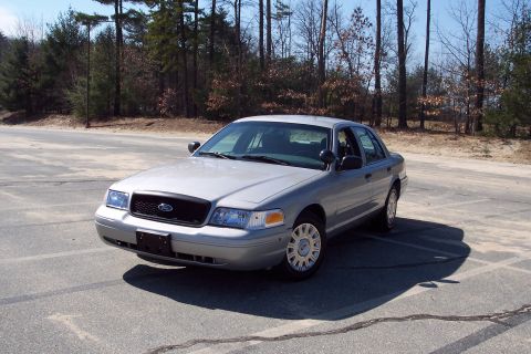 2001 Ford Crown Victoria - A silver 2001 Ford Crown Victoria sedan is parked in an outdoor paved lot with a wooded area in the background.