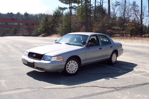 2001 Ford Crown Victoria - A silver 2001 Ford Crown Victoria sedan is parked in an empty asphalt lot with a wooded area and a brick building in the background.