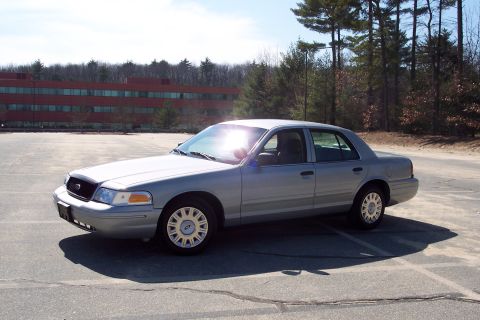 2001 Ford Crown Victoria - A silver 2001 Ford Crown Victoria sedan is parked in a paved lot in front of a modern brick building and a line of trees. The sun is reflec…