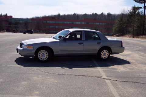 2001 Ford Crown Victoria - A silver 2001 Ford Crown Victoria sedan is parked in a paved lot with a large brick building in the background. The car is viewed from the…