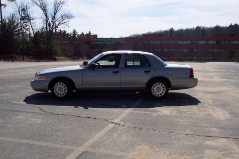 2001 Ford Crown Victoria - A silver 2001 Ford Crown Victoria sedan is parked in an empty asphalt lot with a large brick building in the background.