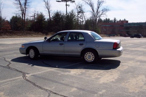 2001 Ford Crown Victoria - The side profile of a silver 2001 Ford Crown Victoria is shown parked in an empty asphalt lot with trees and a building in the background.