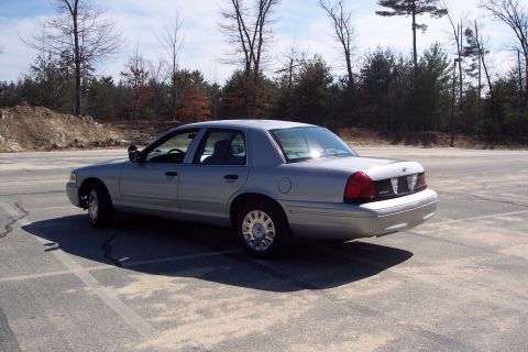 2001 Ford Crown Victoria - A silver 2001 Ford Crown Victoria sedan is parked in an empty asphalt lot with a wooded area in the background. The car is viewed from the…