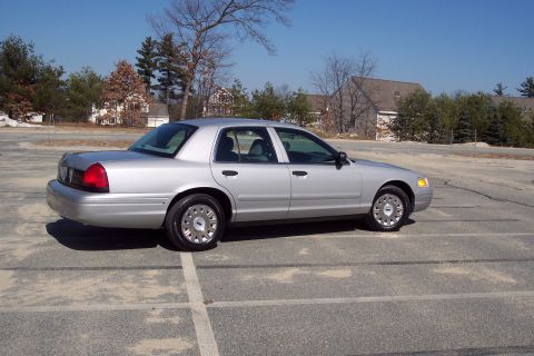 2001 Ford Crown Victoria - A silver 2001 Ford Crown Victoria sedan is parked in an empty lot with buildings and trees in the background.