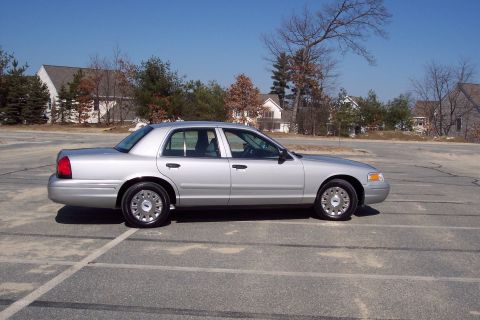 2001 Ford Crown Victoria - A silver 2001 Ford Crown Victoria sedan is parked in an empty asphalt lot with buildings and trees in the background.