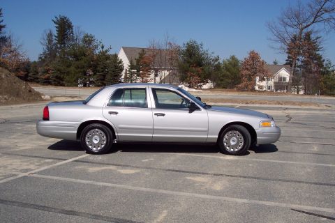 2001 Ford Crown Victoria - A silver 2001 Ford Crown Victoria sedan is parked in an asphalt lot with tire marks. The car is viewed from the side against a backdrop of…