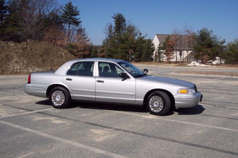 2001 Ford Crown Victoria - A silver 2001 Ford Crown Victoria sedan is parked in an asphalt lot with tire marks. The car is viewed from the side with a wooded area and…