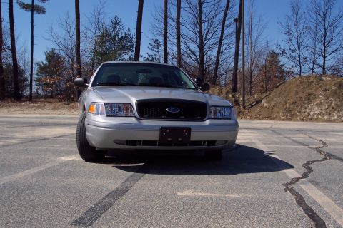 2001 Ford Crown Victoria - The front view of a silver 2001 Ford Crown Victoria parked on asphalt with trees and a dirt pile in the background.