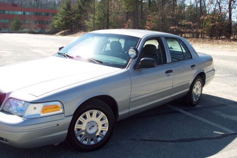 2001 Ford Crown Victoria - The front passenger side of a silver 2001 Ford Crown Victoria is shown parked in an asphalt lot with trees and a building in the background…