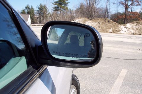 2001 Ford Crown Victoria - Close-up view of a side mirror on a white vehicle, with a blurred background of trees and sky.