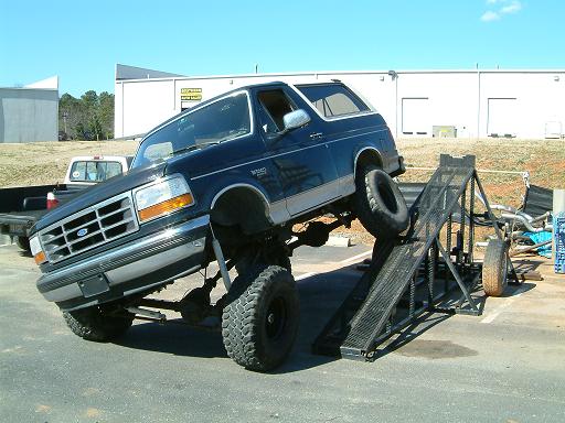 Lifted dark blue Ford Bronco flexing front wheel up a black steel ramp in a paved lot