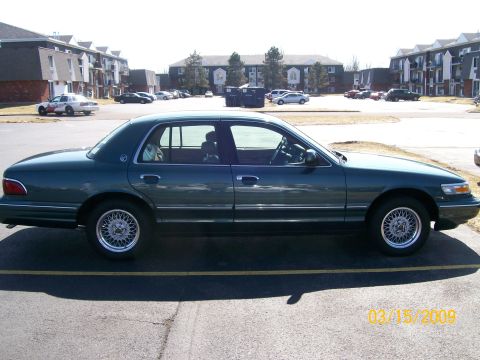 1995 Mercury Grand Marquis - A dark green 1995 Mercury Grand Marquis sedan is seen from the side in a parking lot with apartment buildings in the background.