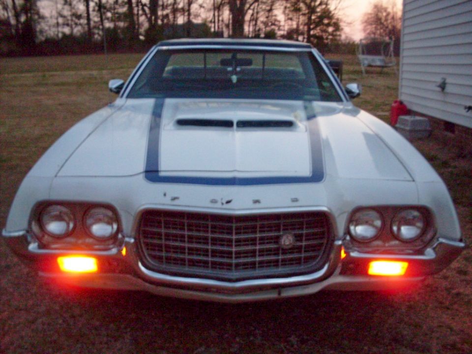 Front of white Ford with blue stripes, quad headlights, lit turn signals, parked at dusk.