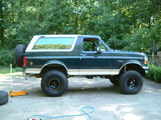 Lifted dark green two-tone Ford Bronco with big off-road tires, side view on a driveway among trees